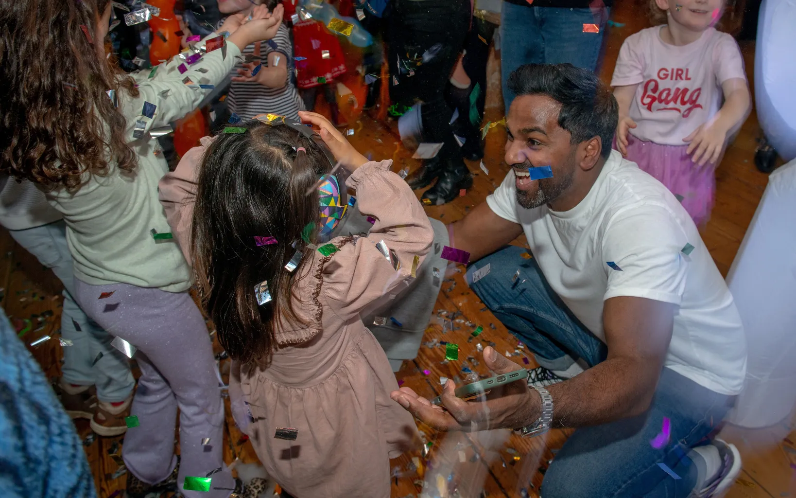 Adults and children enjoy a lively indoor party with colorful confetti in the air. A man crouches to talk to a child amid the festivities.