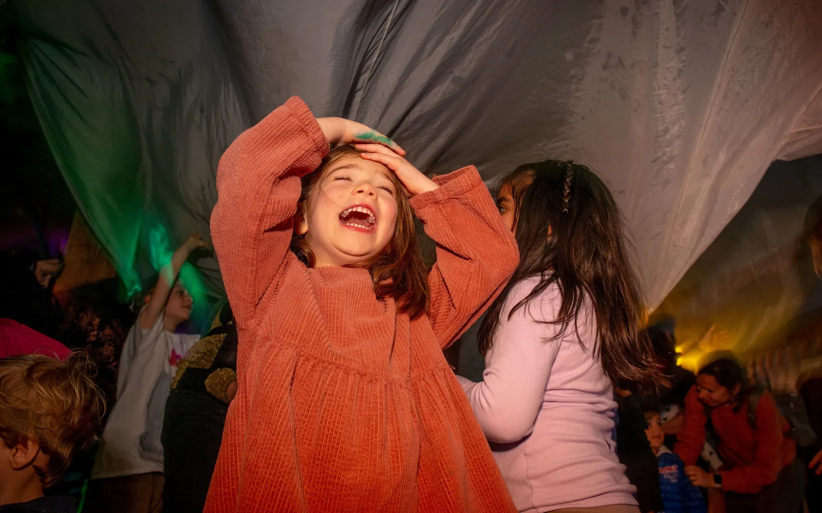Children are laughing and playing under a large fabric sheet, with colorful lights in the background.