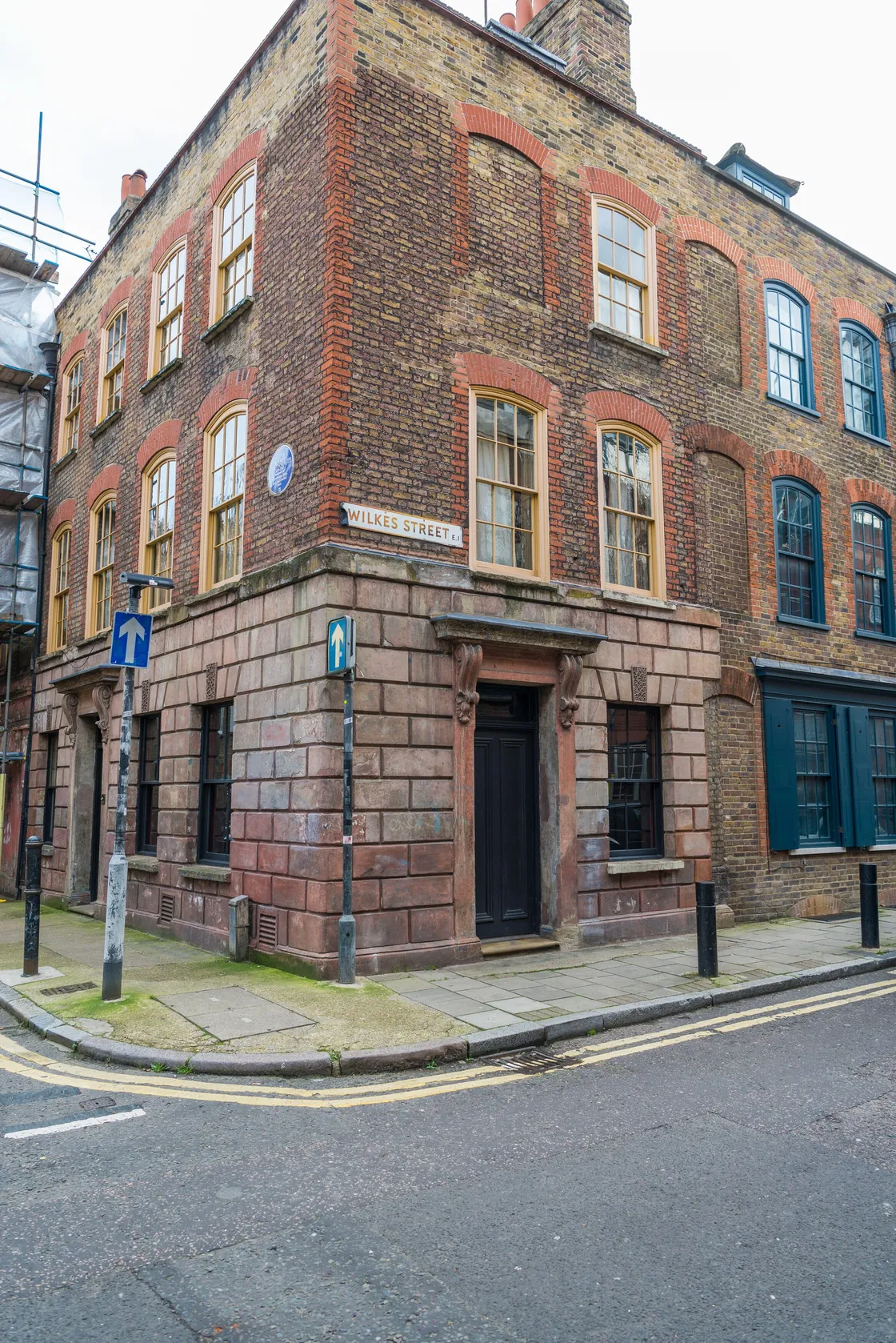 A London townhouse on the corner of a street with a blue plaque next to one of the windows, and with a street sign reading 'Wilkes Street E1'