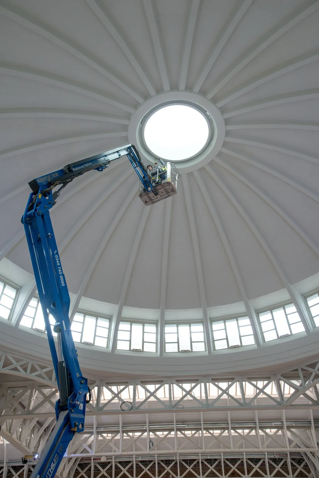 A worker in a blue lift performs maintenance near the central skylight of a large domed ceiling, with radial beams and windows around the base, high above the floor of this impressive London venue.