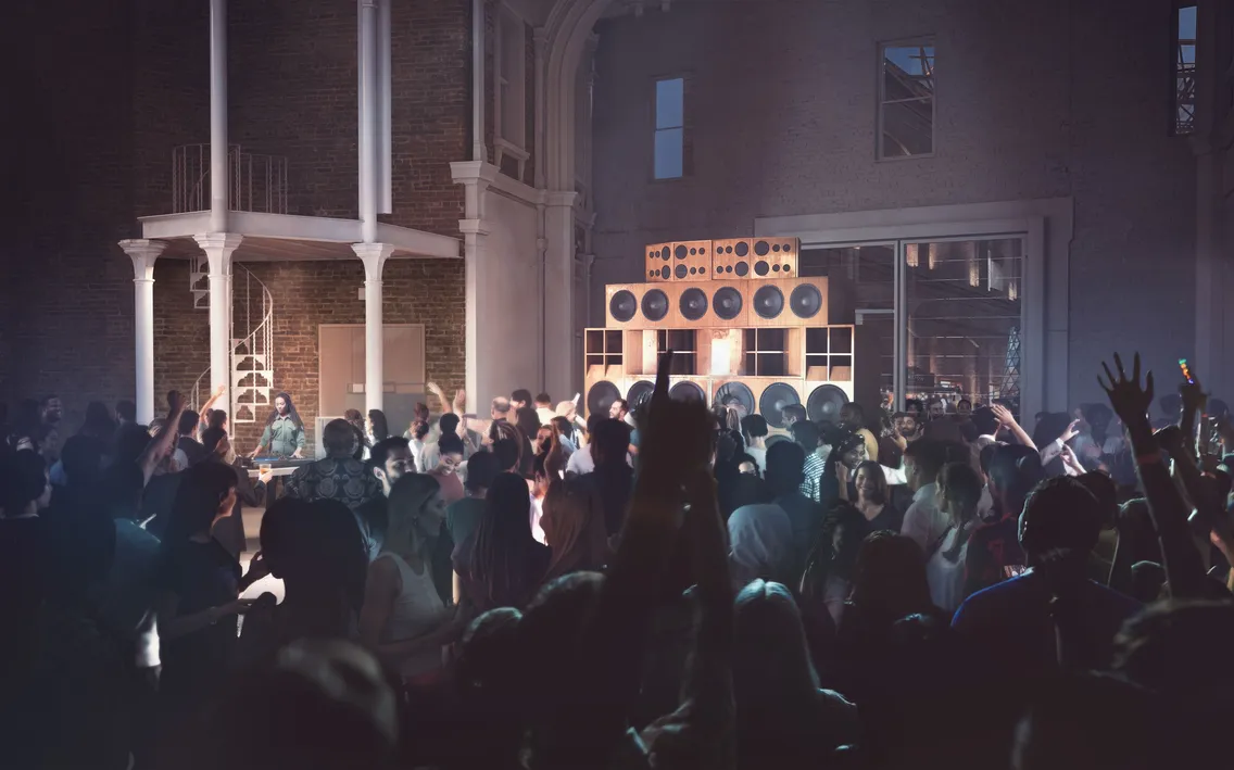 A large crowd dances in a dimly lit indoor venue with tall speakers stacked near a brick wall, as the spiral staircase in the background overlooks the lively market atmosphere.