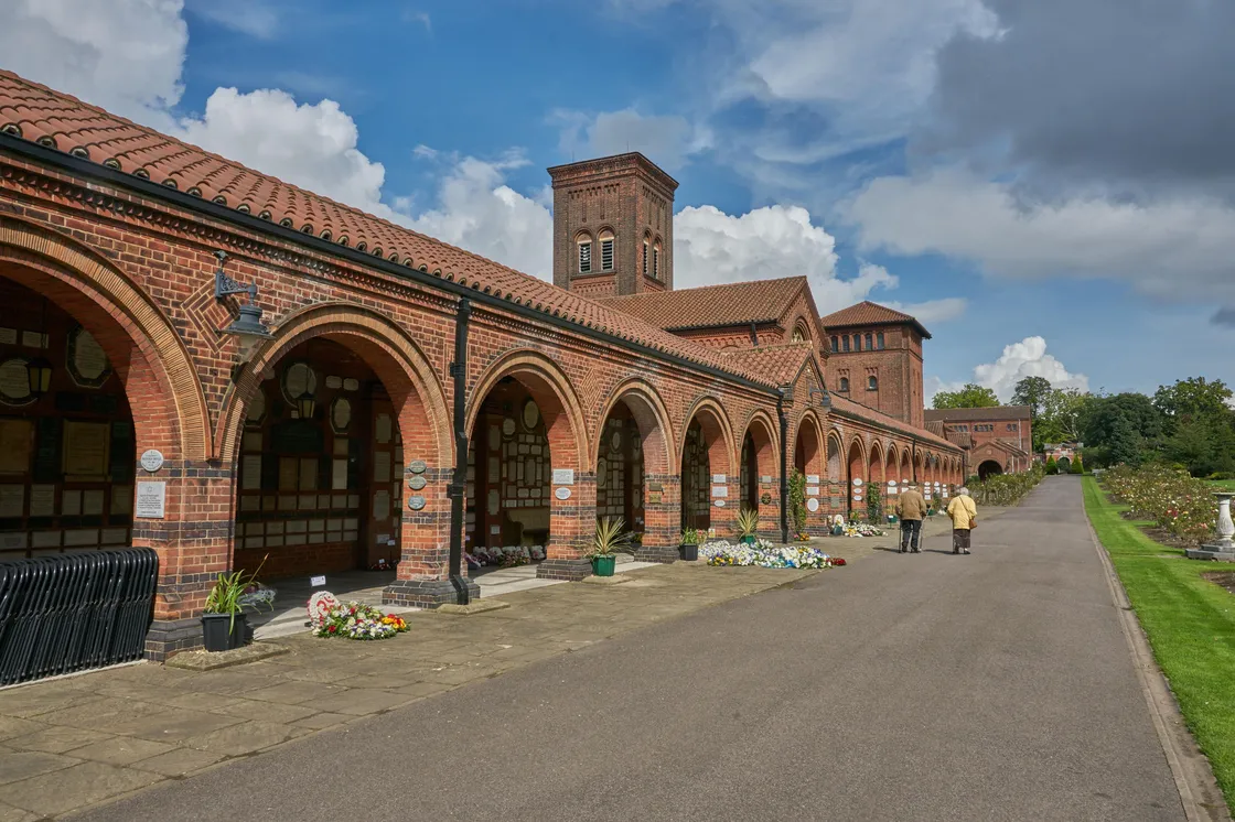 A brick columbarium with arched walkways, memorial plaques, and flower arrangements, with a few people walking along the paved path beside it.