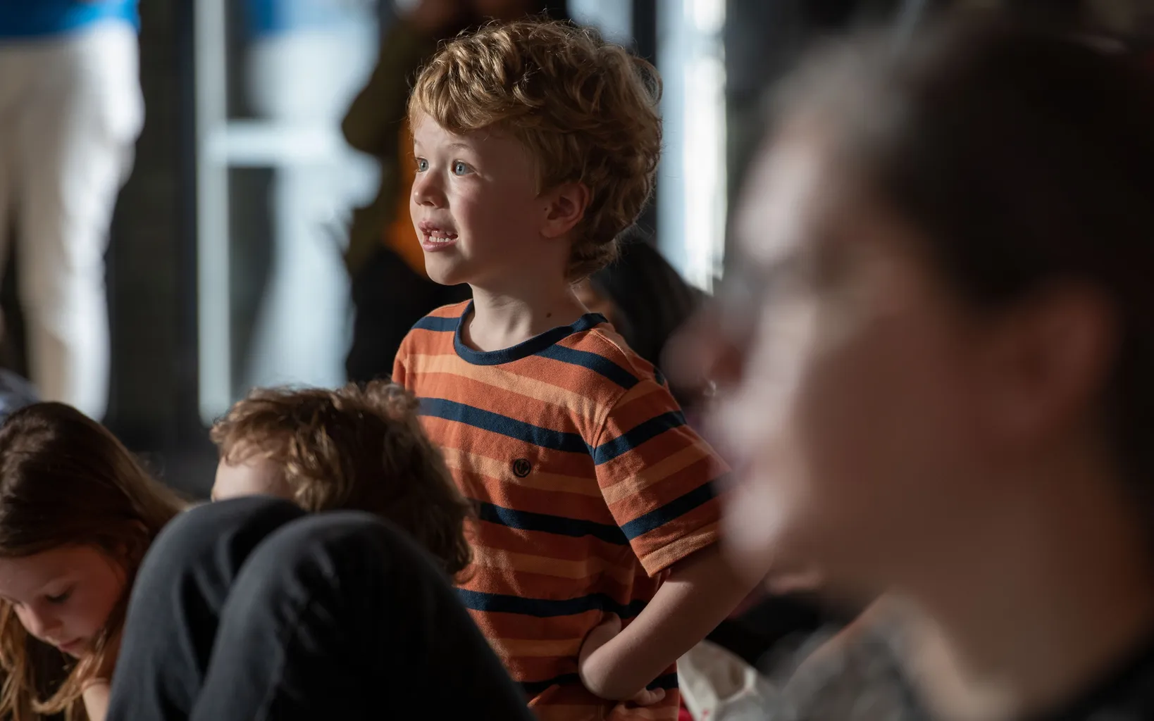 A young boy in a striped orange shirt looks ahead with interest, surrounded by other children in a softly lit indoor setting.