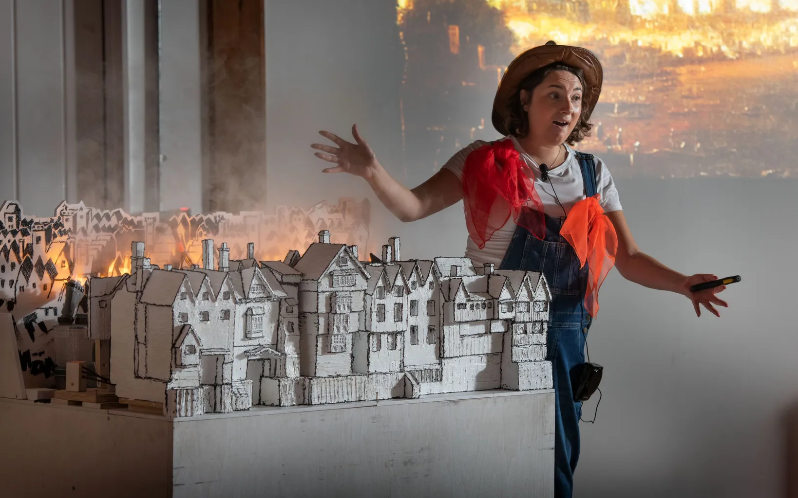 A woman in costume gestures while presenting a model of historic buildings, with smoke and a burning city scene projected in the background.