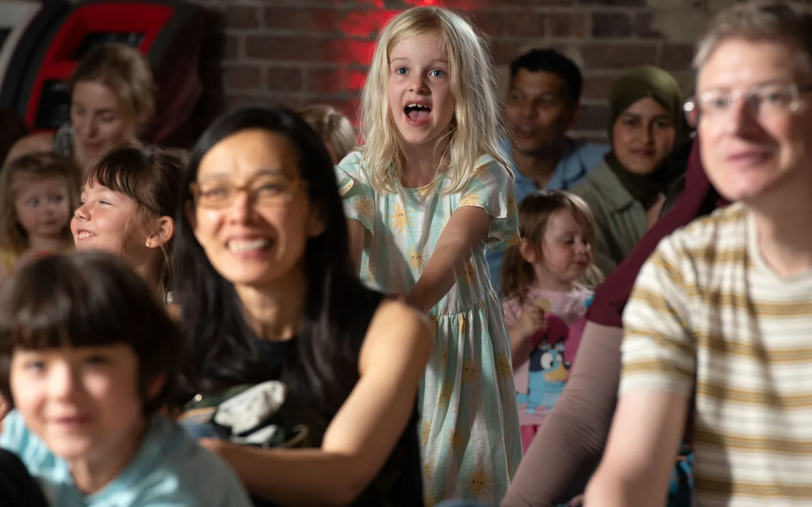 An audience sits on the ground, with various people smiling, though most are blurred. The focus of the image is a small blonde girl who stands in the centre of the picture, wearing a mint green and white striped sun dress. Her mouth is open.