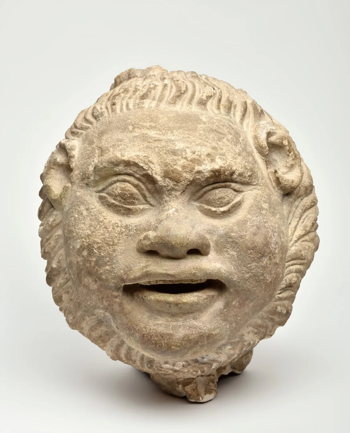 Stone sculpture of a grotesque face with wide-open mouth and curly hair, featuring expressive eyes and a textured surface.