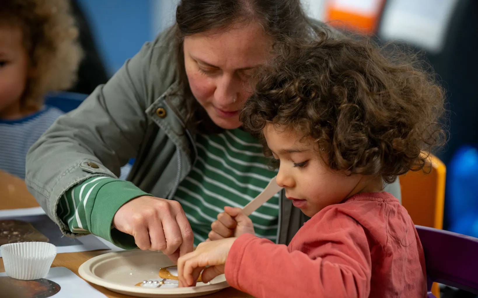 An adult and a child sit at a table, working together to decorate cookies on a paper plate using small utensils.