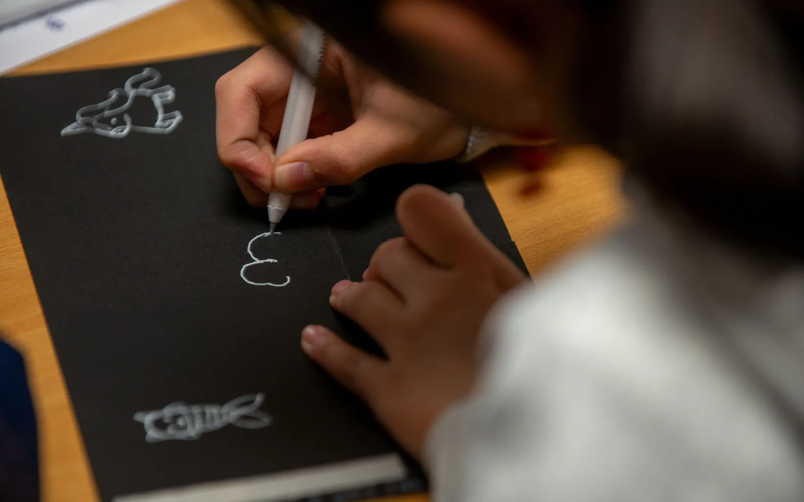 A child draws white animal shapes and letters on black paper using a white pen, with focus on their hands and artwork.