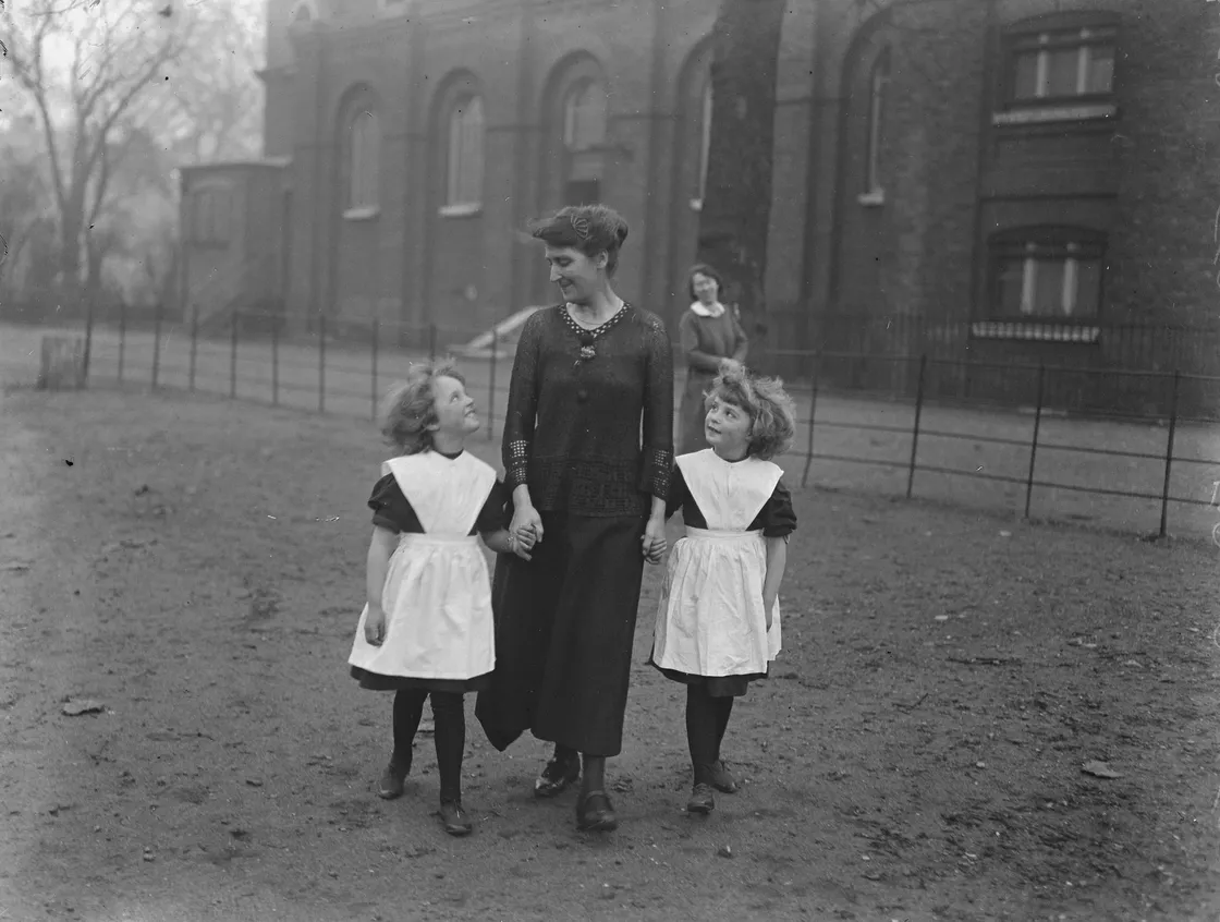 A woman walks outside holding hands with two young girls in matching dresses and aprons, with a building and another person in the background.
