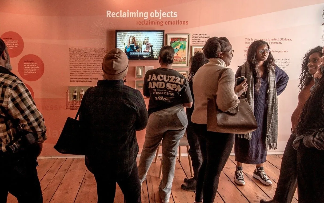 A group of people stand in front of a museum exhibit titled "Reclaiming objects," reading text and watching a video display on a wall.