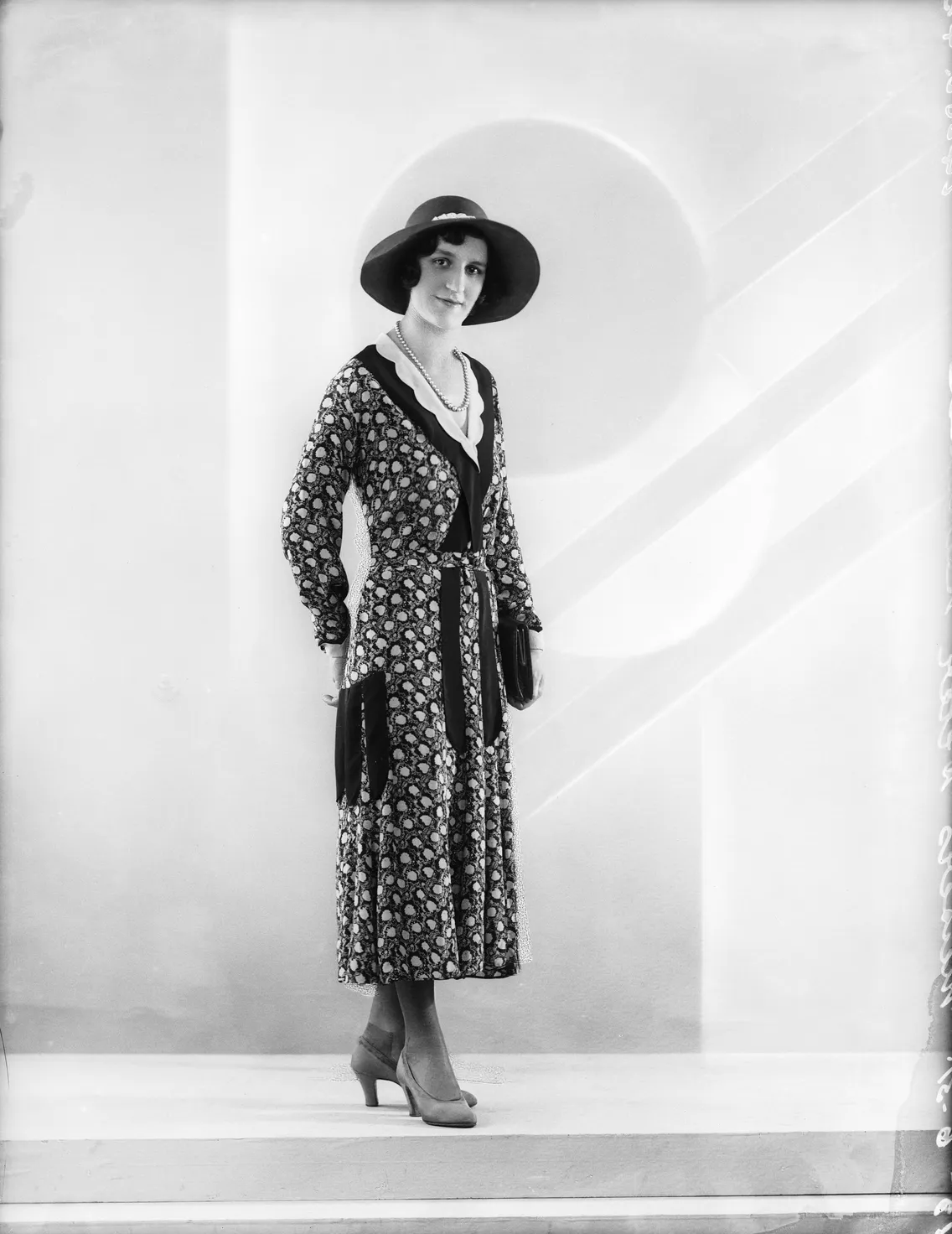 A black and white photo of a woman standing in a floral dress, wide-brimmed hat, and heels, posing with a subtle smile.