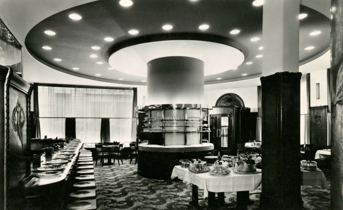 Black-and-white photo of the vintage restaurant Prunier, with round ceiling lights, ornate woodwork, set tables, and a central circular buffet area providing a striking view of classic elegance.