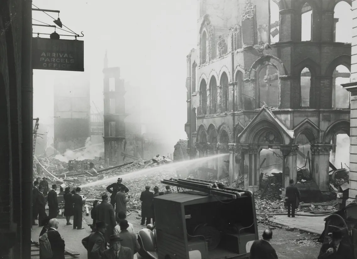 Firefighters spray water on rubble after bombing damage in a city, with onlookers and a partially destroyed building visible.