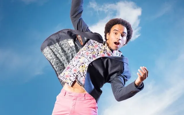 A man in a floral shirt, blazer, and pink pants appears to jump or float in the air against a blue sky with clouds in this striking photo reminiscent of a Chris Close portrait.