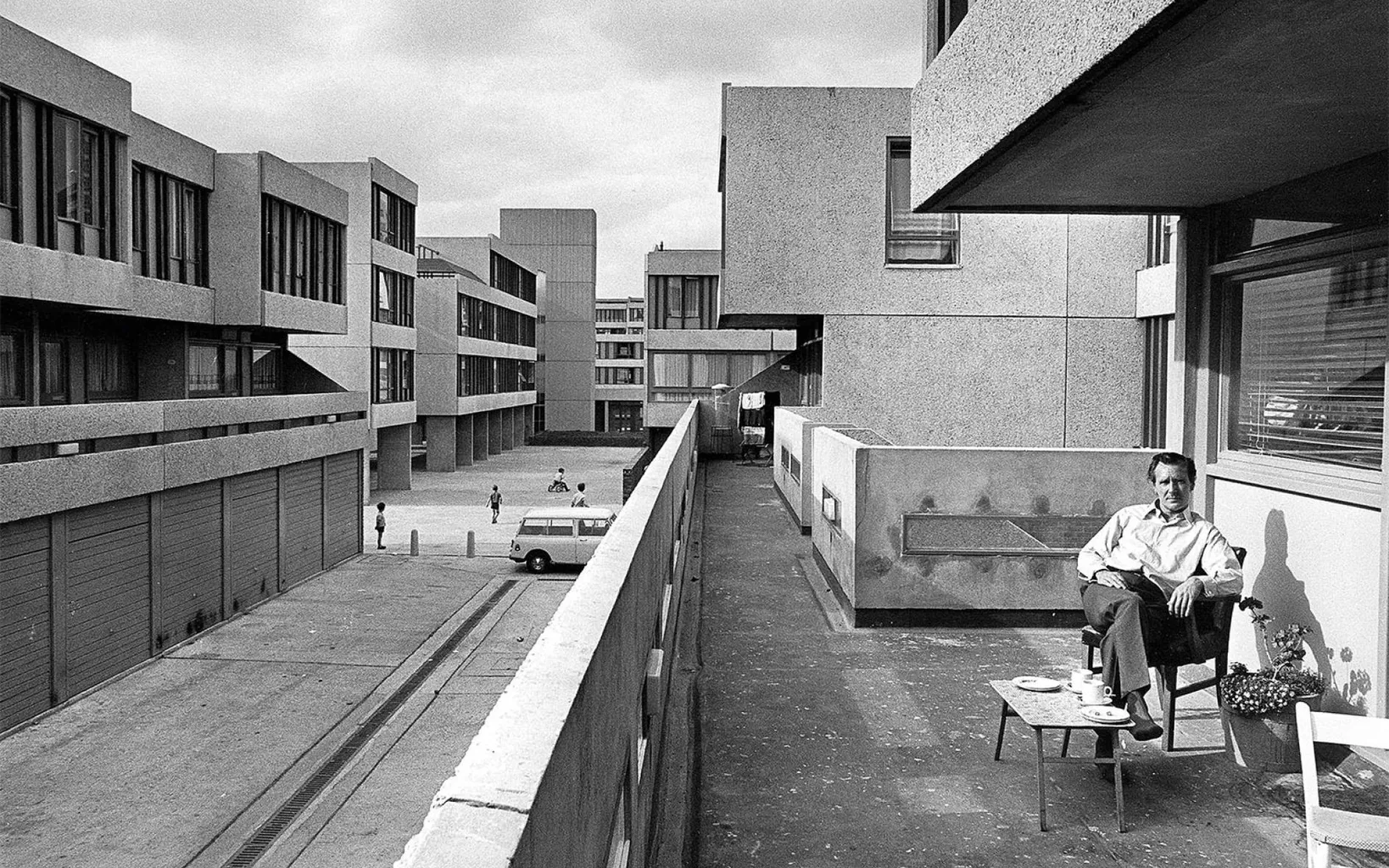 A man sits on a balcony with a table and chair, overlooking a concrete housing estate with garages, parked car, and a few people walking below.