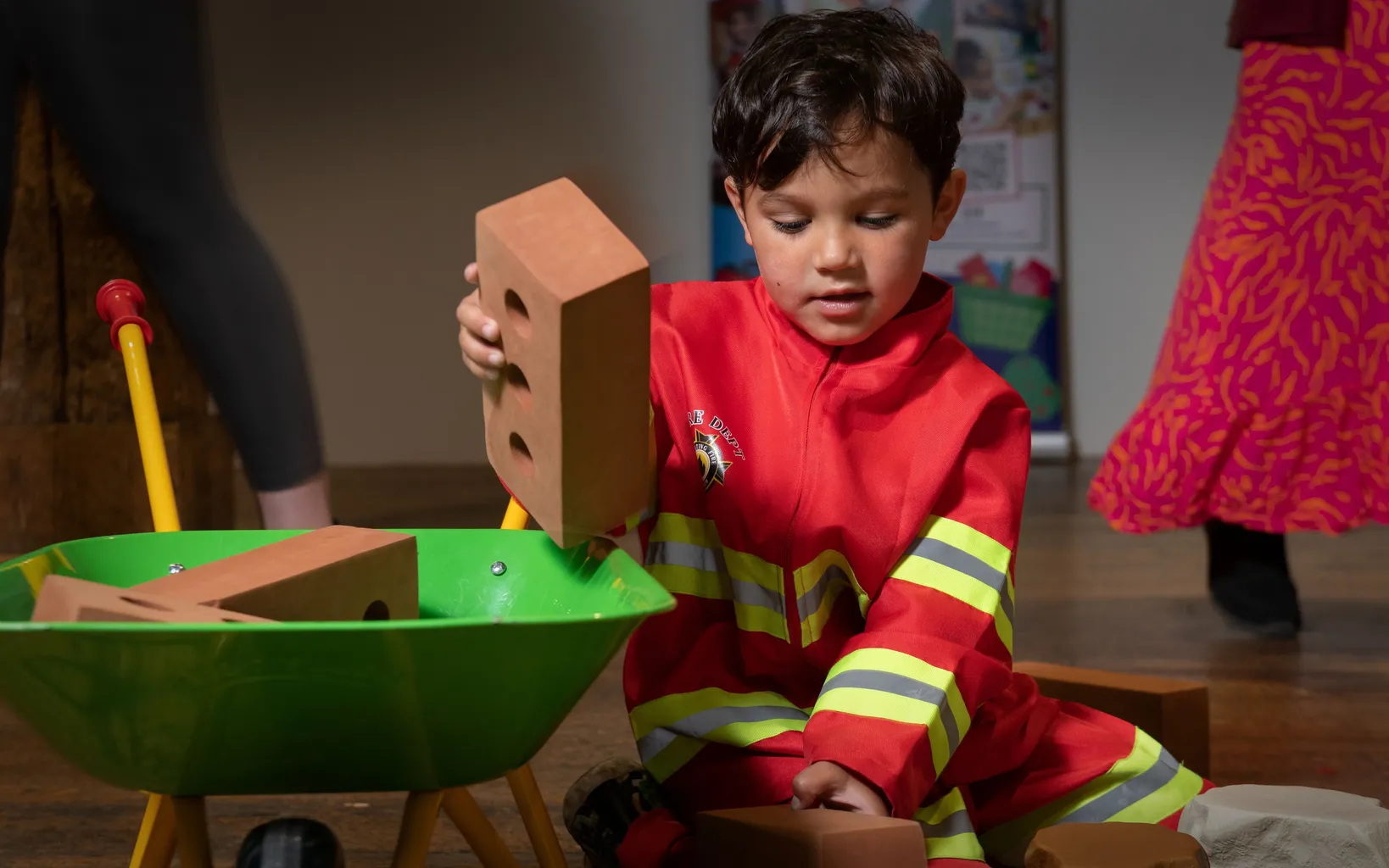 A young boy is sitting on a wooden floor in a high vis jacket. Next to him in a small green wheelbarrow that he is using to load toy bricks into.