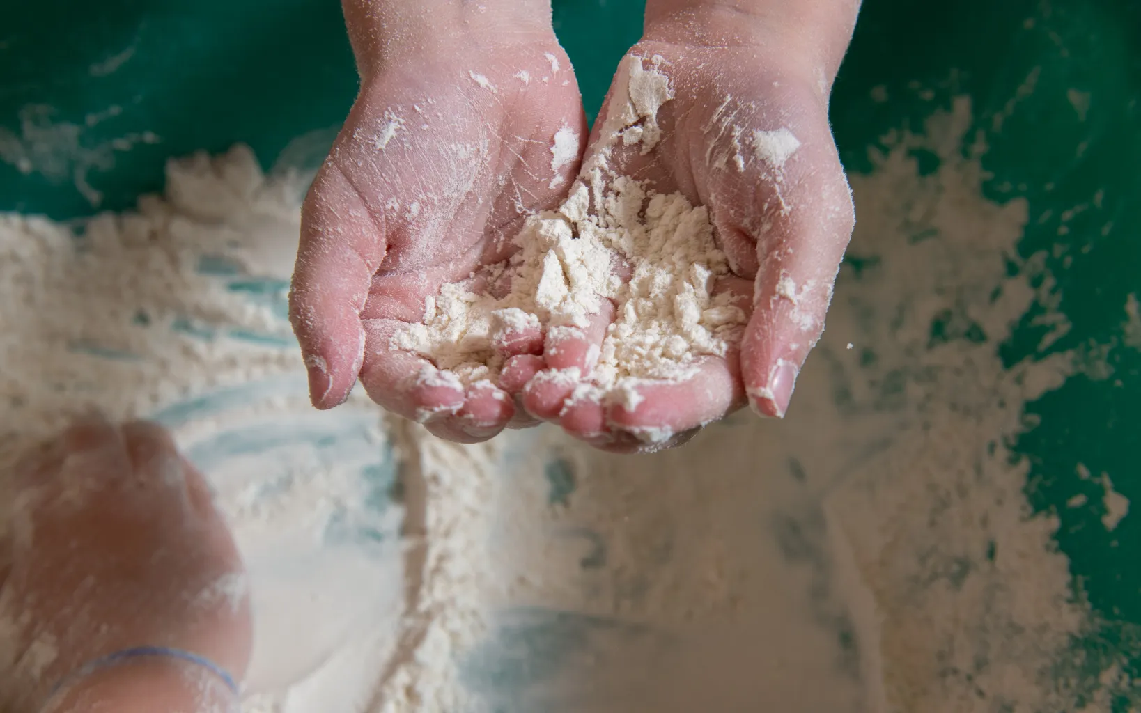 A child's hands hold a wooden rolling pin over a flattened circle of clay.
