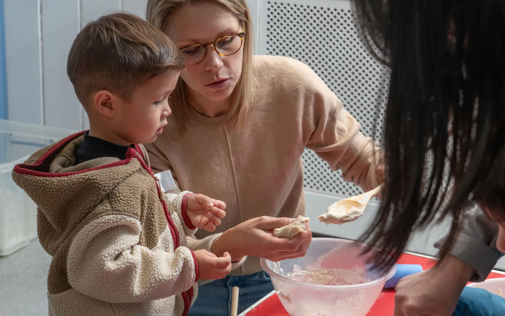 An adult and a young child mix dough in a white bowl with wooden spoons, sitting at a table with a blue tinsel backdrop behind them.