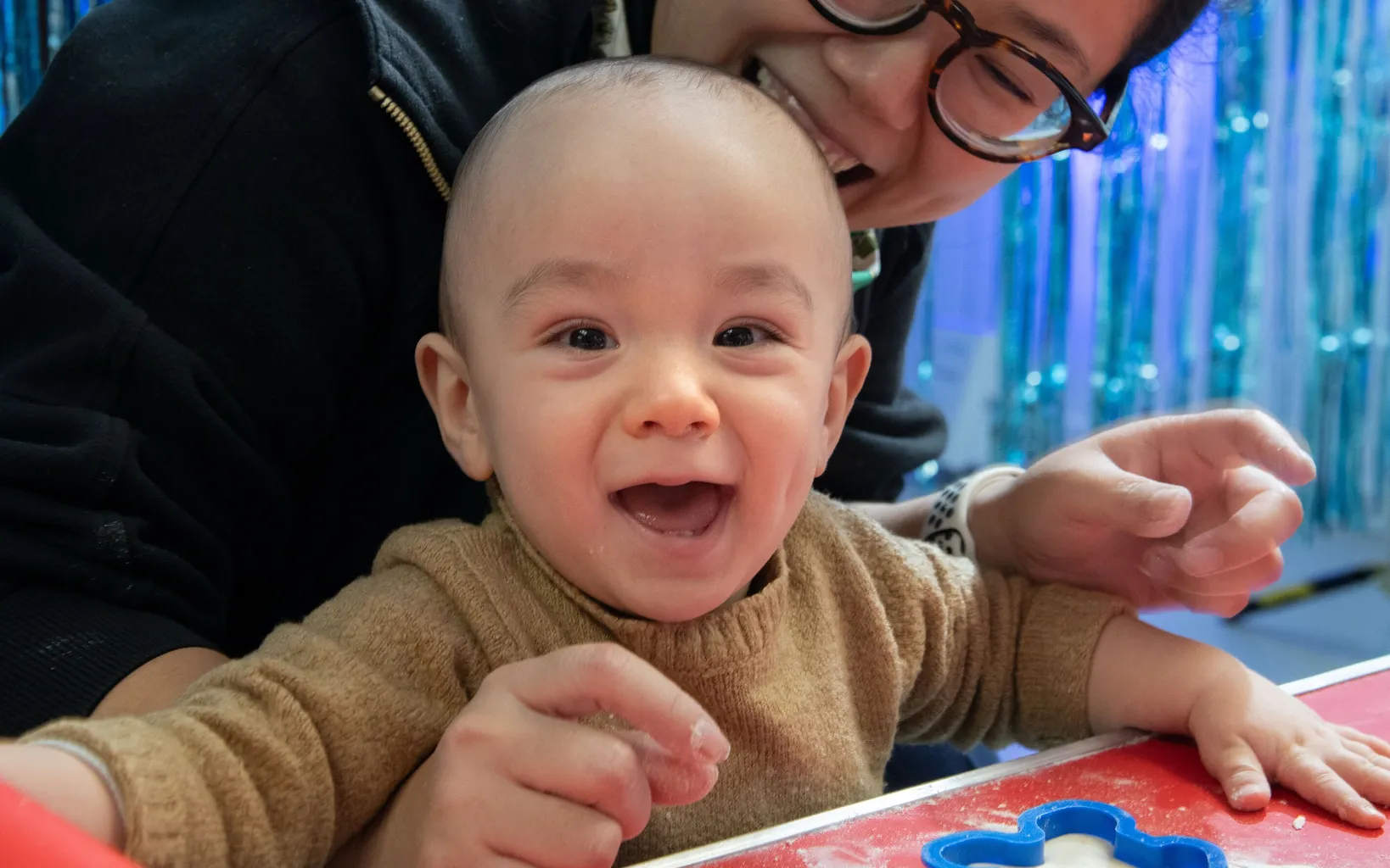 An adult and a baby smile at a table with baking supplies, including a rolling pin and cloud-shaped cookie cutter, with blue streamers in the background.