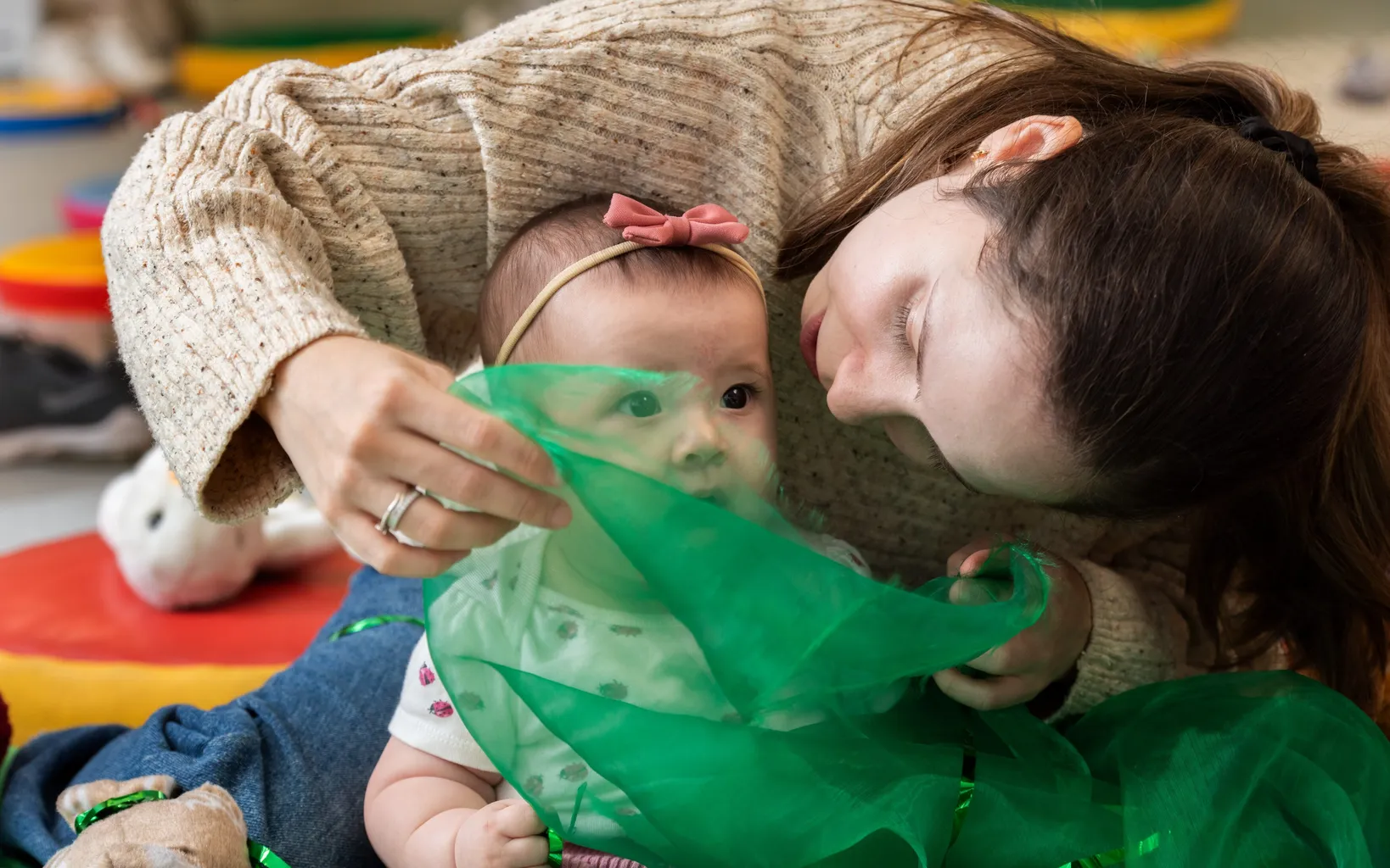 A woman sits on the floor with a baby on her lap. The woman is bending over the baby and waving a sheer green scarf in her face.