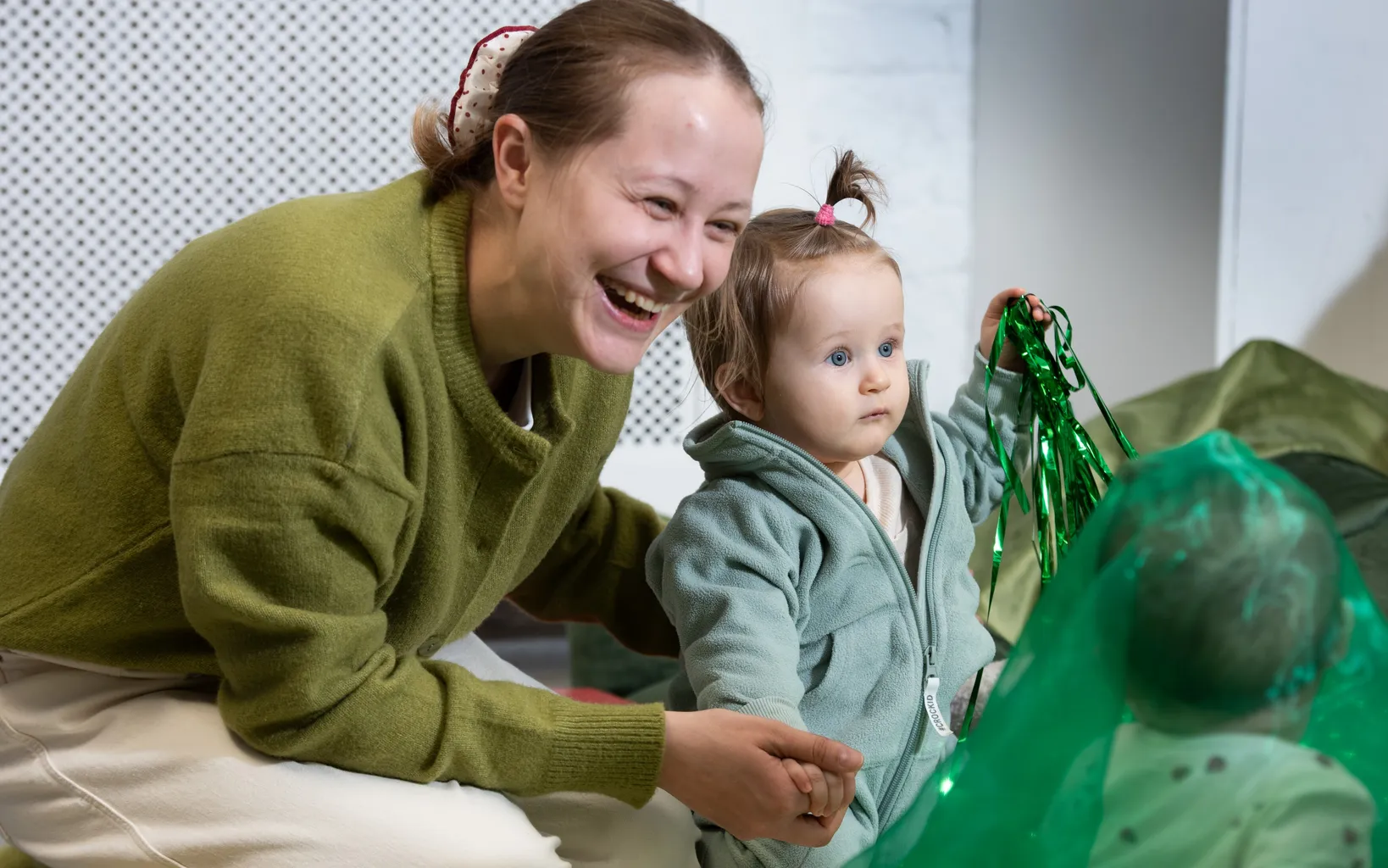 An adult and a toddler sit on the floor; the adult smiles while the toddler holds shiny green ribbon and looks ahead.