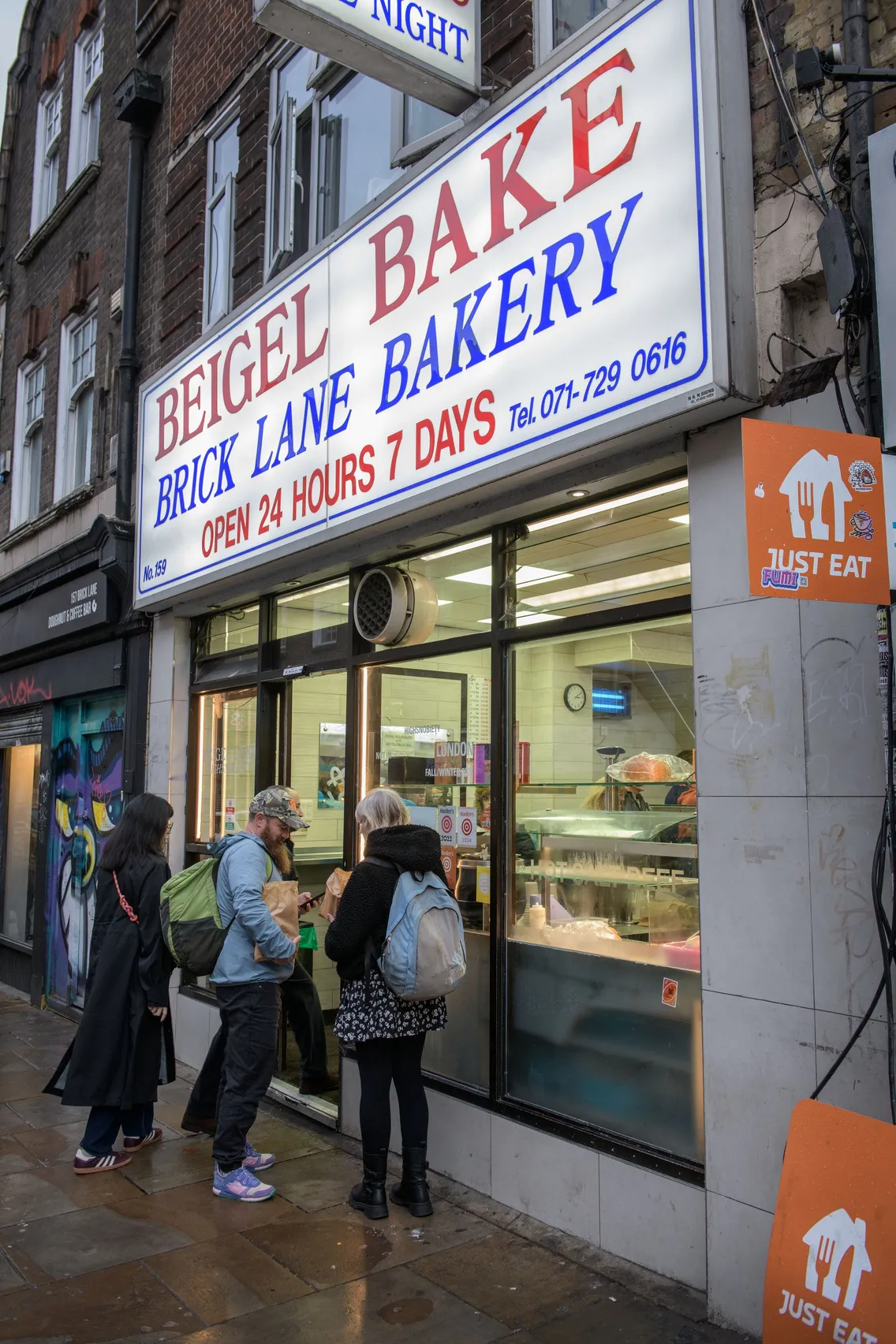 Three people stand outside Beigel Bake Brick Lane Bakery, a 24-hour shop, looking at the menu through the window on a wet sidewalk.