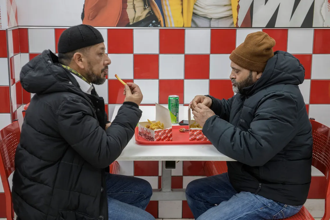 Two men in winter jackets and hats sit at a small table in a checkered restaurant, eating fries and burgers with a can of soda between them.