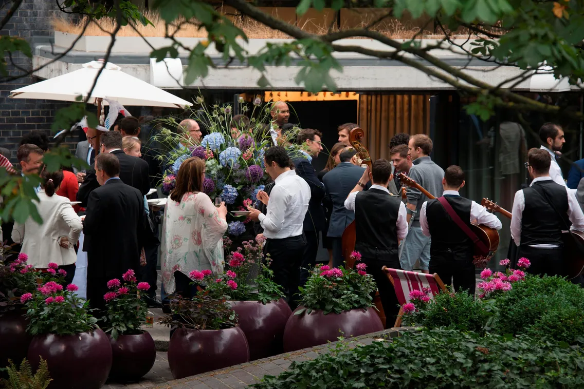 A group of people socializing at an outdoor event, surrounded by potted plants and musicians playing string instruments in the background.
