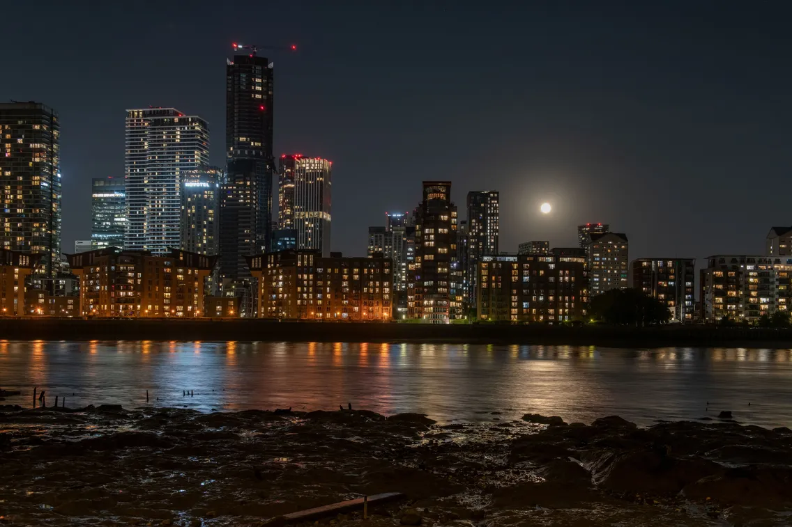 City skyline at night with illuminated buildings reflecting on the water; a bright full moon is visible in the sky.