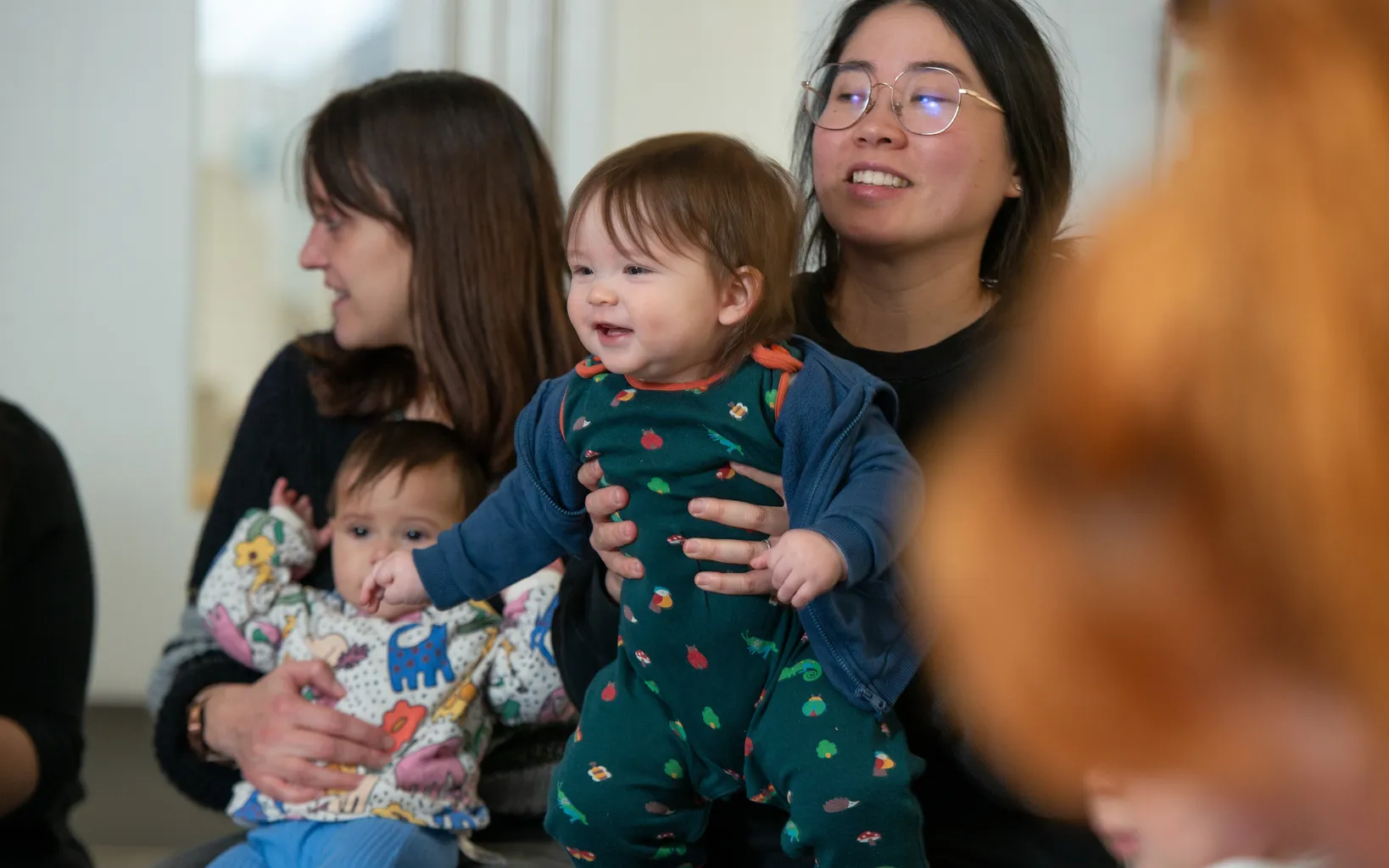 Two women are sitting, each holding a baby. One woman holds a baby with printed pajamas, and the other holds a baby in a patterned outfit. The background is an indoor setting.