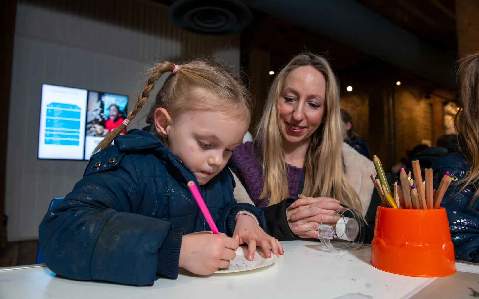 A young girl writes with a pink marker at a table while a woman looks on, smiling. Pencils are placed in an orange holder nearby.
