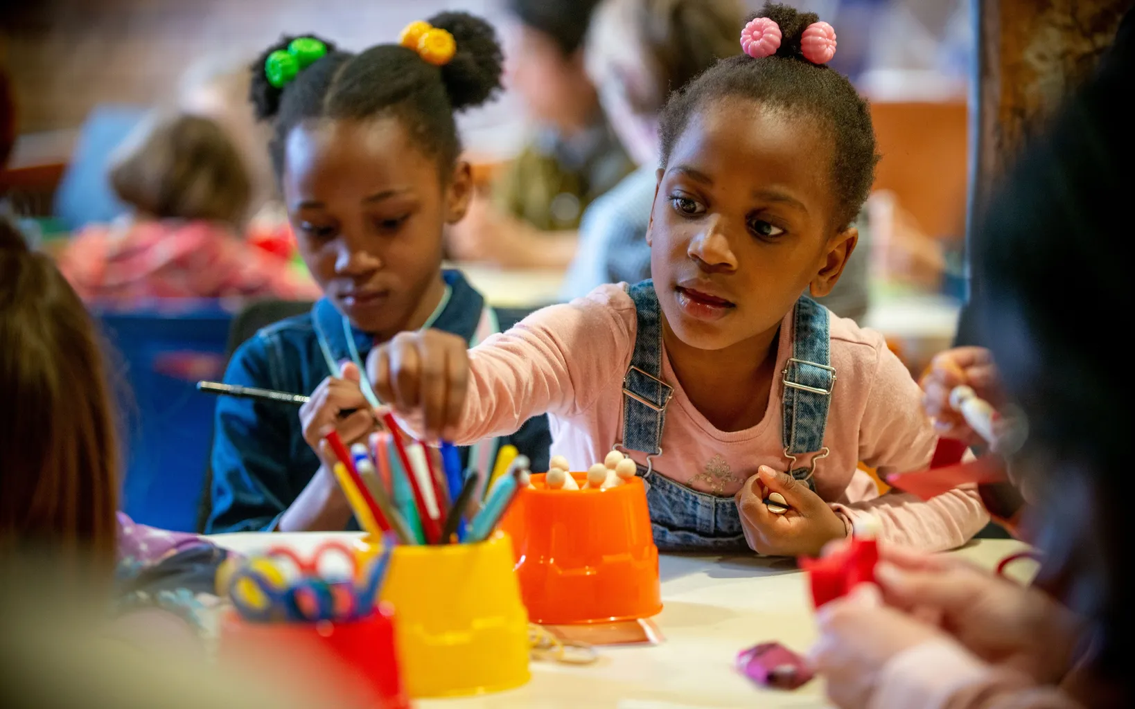 Two young girls sit at a wooden table with craft supplies. One is colouring as another reaches for colouring pencils.
