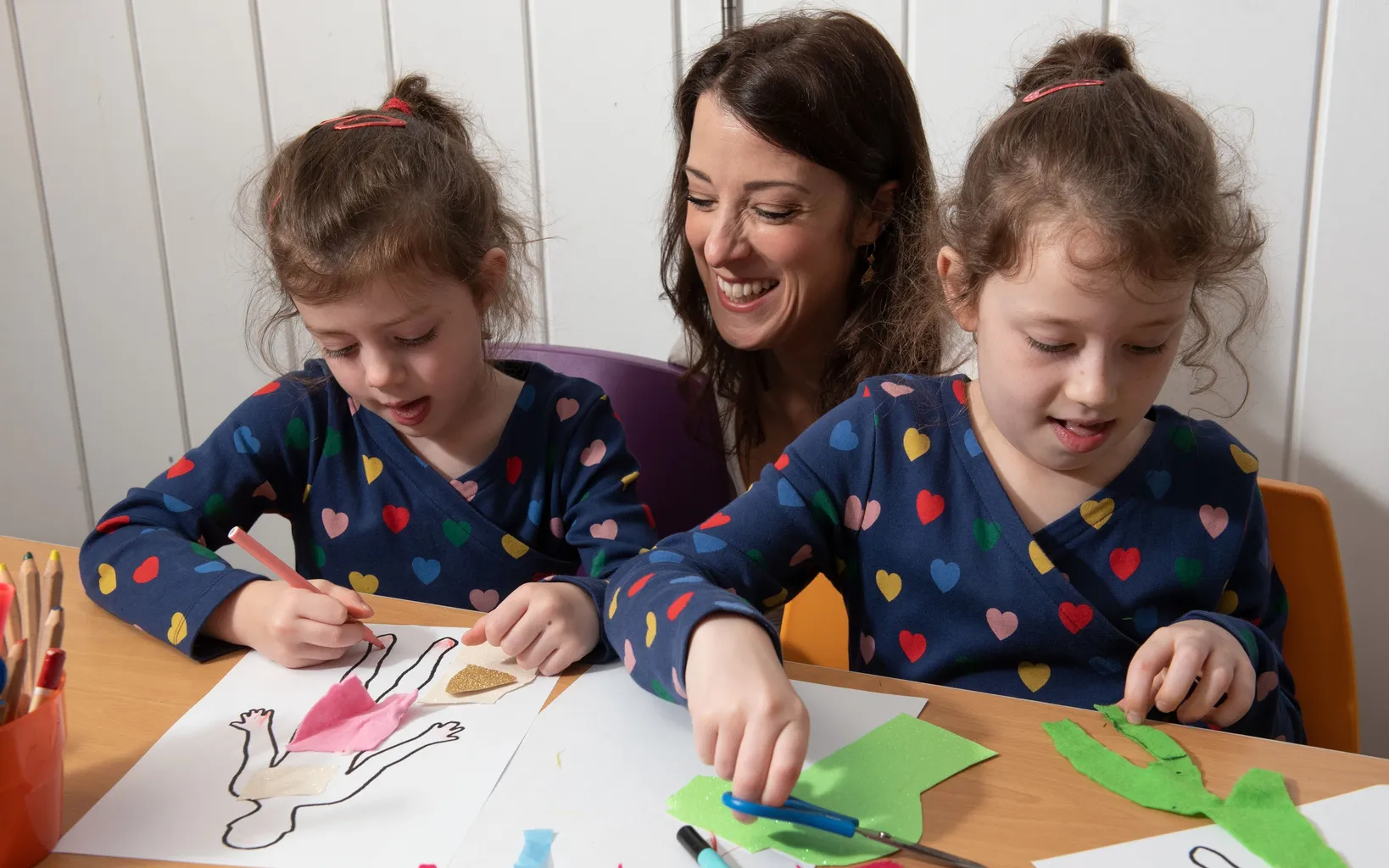 Two young girls in heart-patterned outfits craft on paper with colored felt, guided by a woman behind them, smiling.