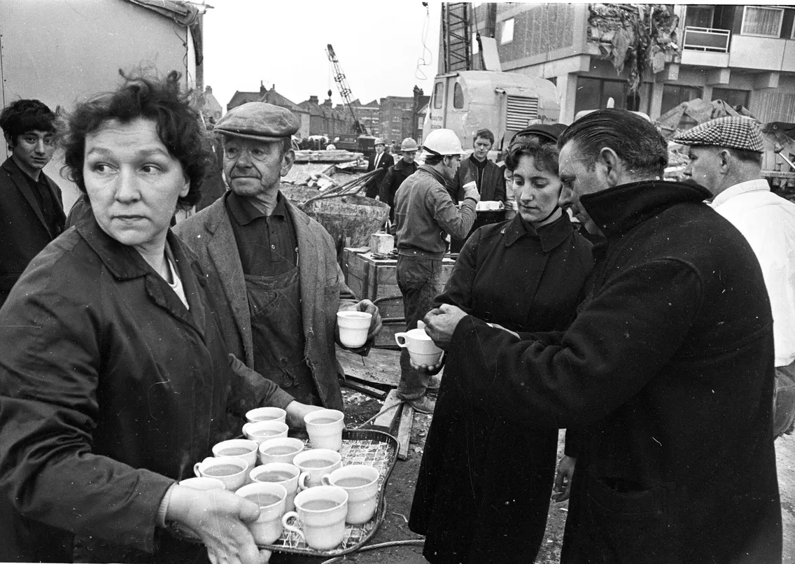 A busy scene with emergency workers in the background and a handful of people in the foreground, with one woman holding ma tray of tea