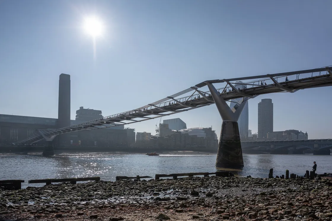 Millennium Bridge in London spans the River Thames, with the Tate Modern and city skyline visible. The sun shines brightly in a clear sky.