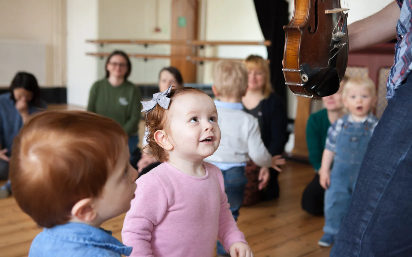 Two toddlers stand in the foreground, smiling and staring at a held violin. Other families sit a on a wooden floor behind them.