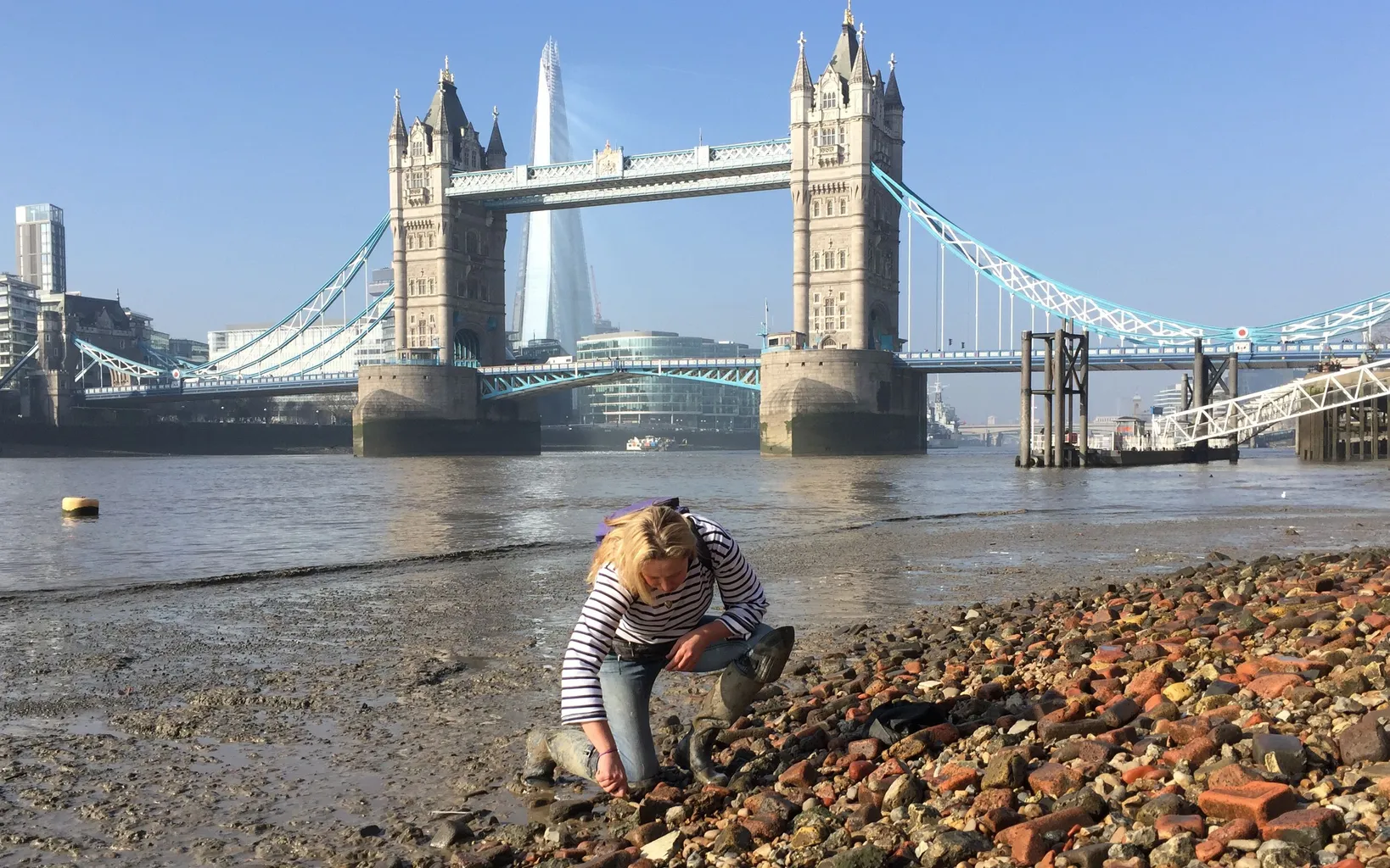 A woman in boots stands on rocks in shallow water with the O2 Arena and city buildings in the background.