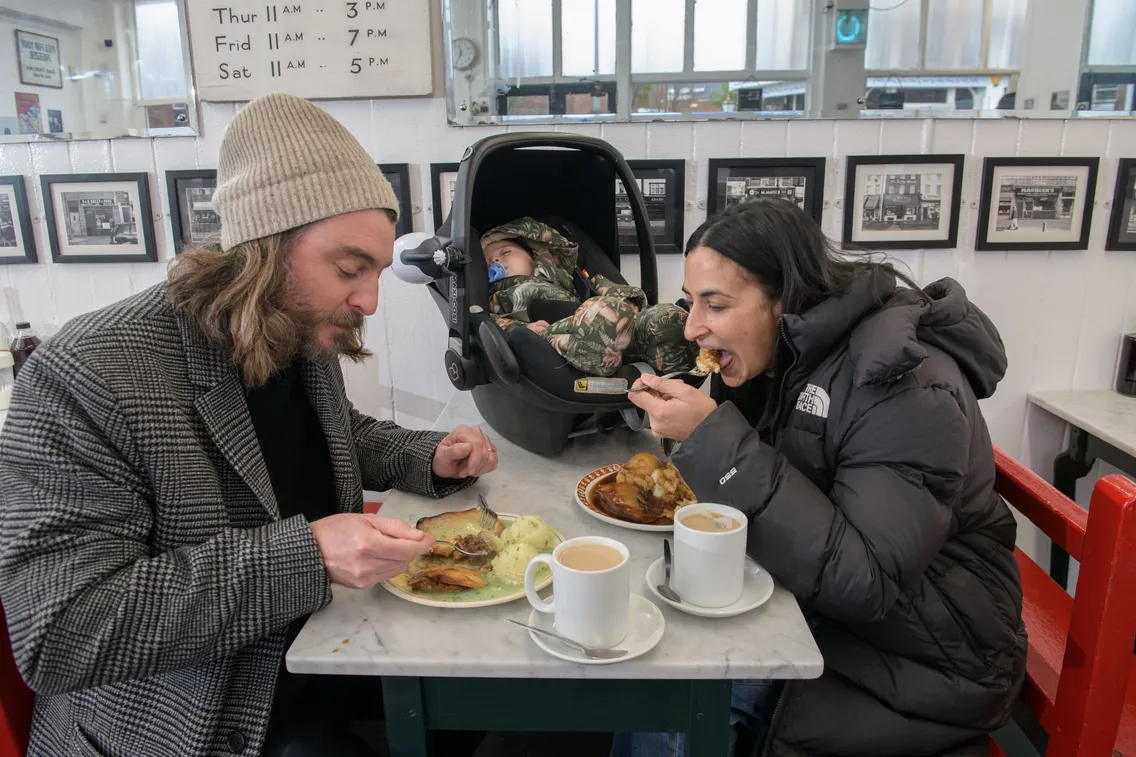 Two adults eat a meal at a small table in a café while a baby in a car seat sleeps beside them; two mugs of tea are on the table.