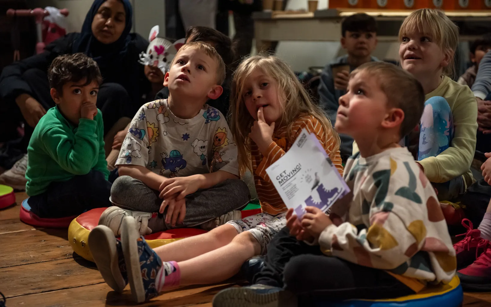 A group of children sat on the floor looking upwards at something out of shot.