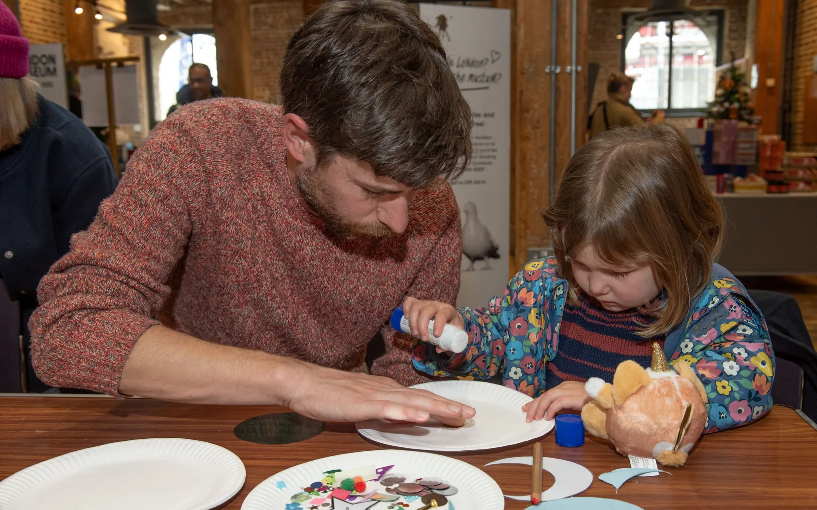 A man sits at a table with a small girl. Both of their heads are bent down as they stick shapes onto a paper plate.