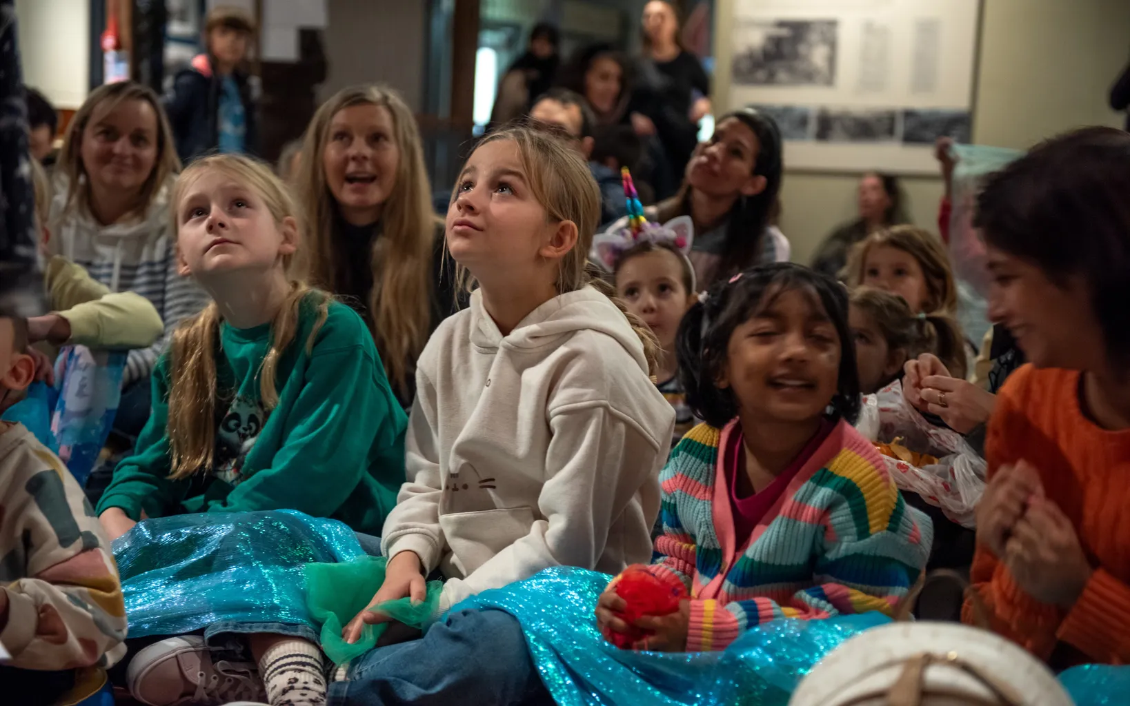 A group of children are sitting on a wooden floor. Their faces are tilted upwards, watching something happening off-camera. A length of blue shimmery fabric is laying across their laps.