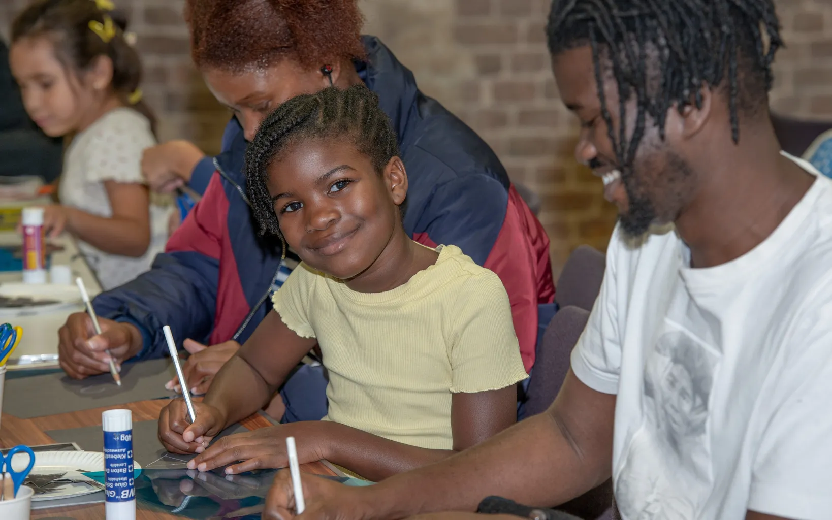 A young girl sits at a table, smiling while drawing with a marker. Adults and children are engaged in art activities around her in a room with brick walls.