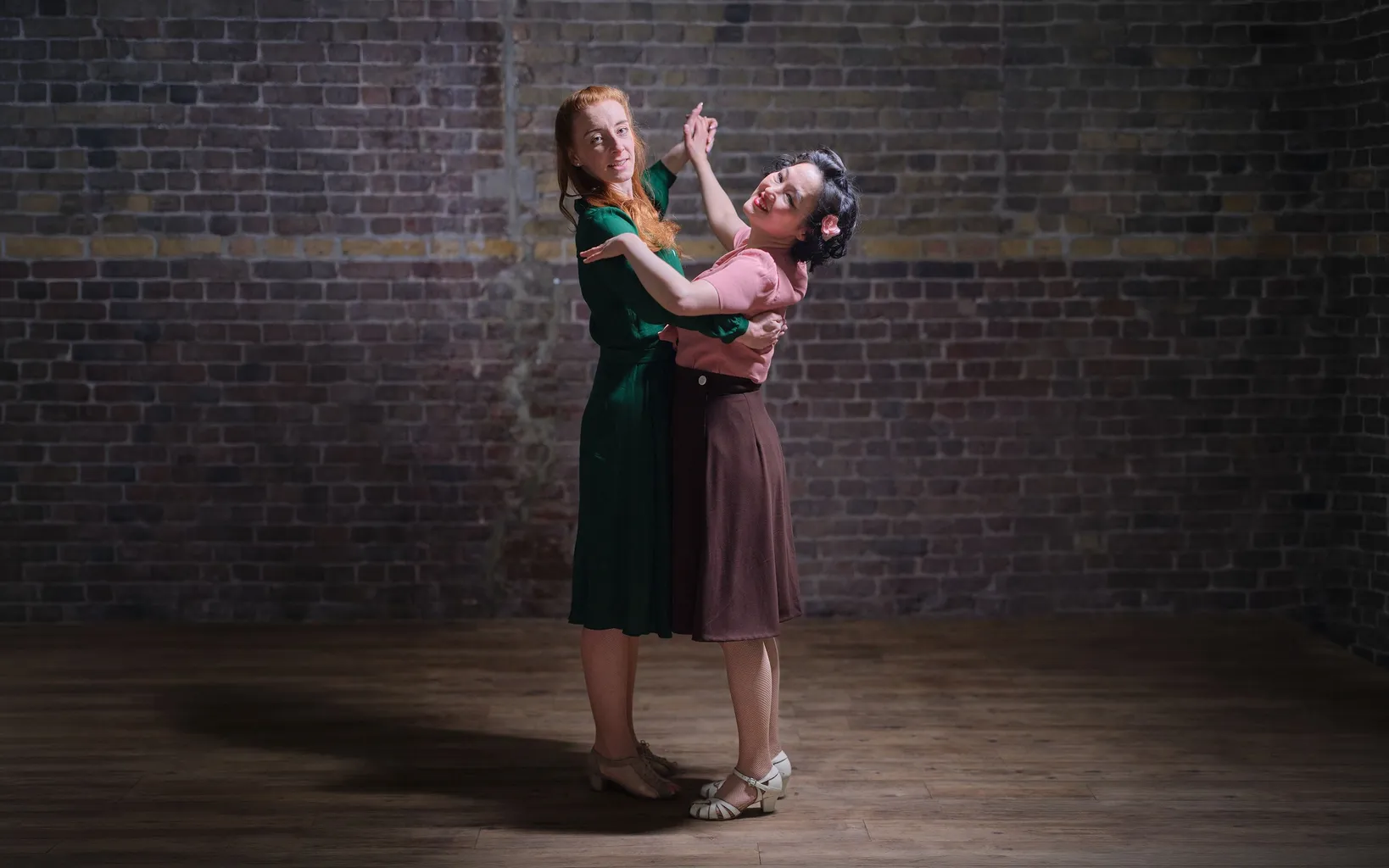 Two women in vintage-style dresses dance together on a wooden floor in front of a brick wall under focused lighting.