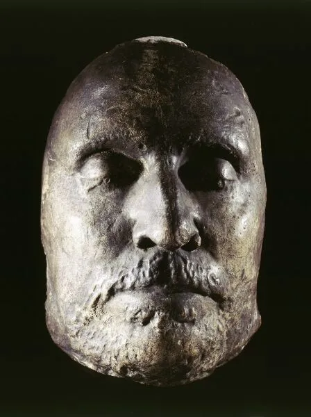 Ancient bronze sculpted head with detailed facial features, including a beard and closed eyes, against a dark background.
