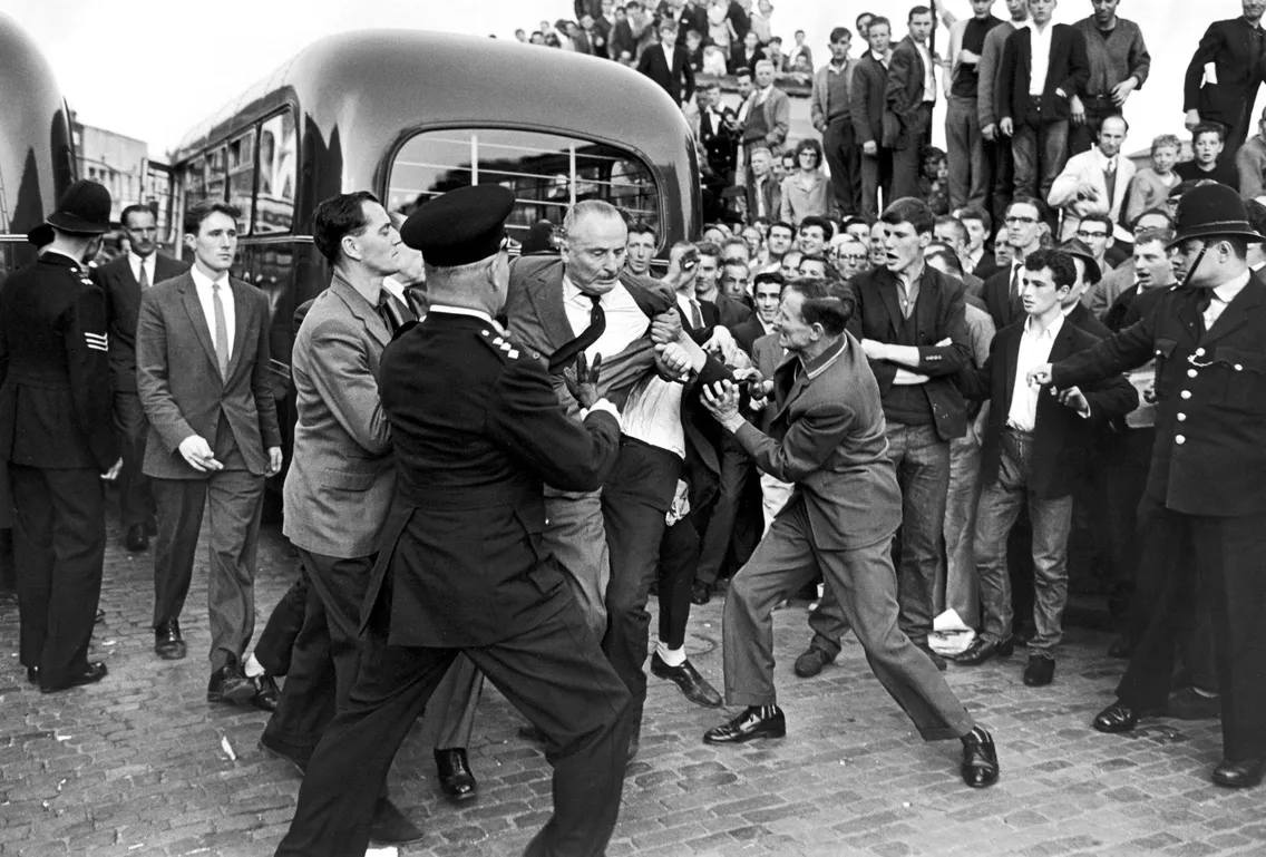 A busy crowd of people around a bus, with a man being grabbed by a handful of men in the centre