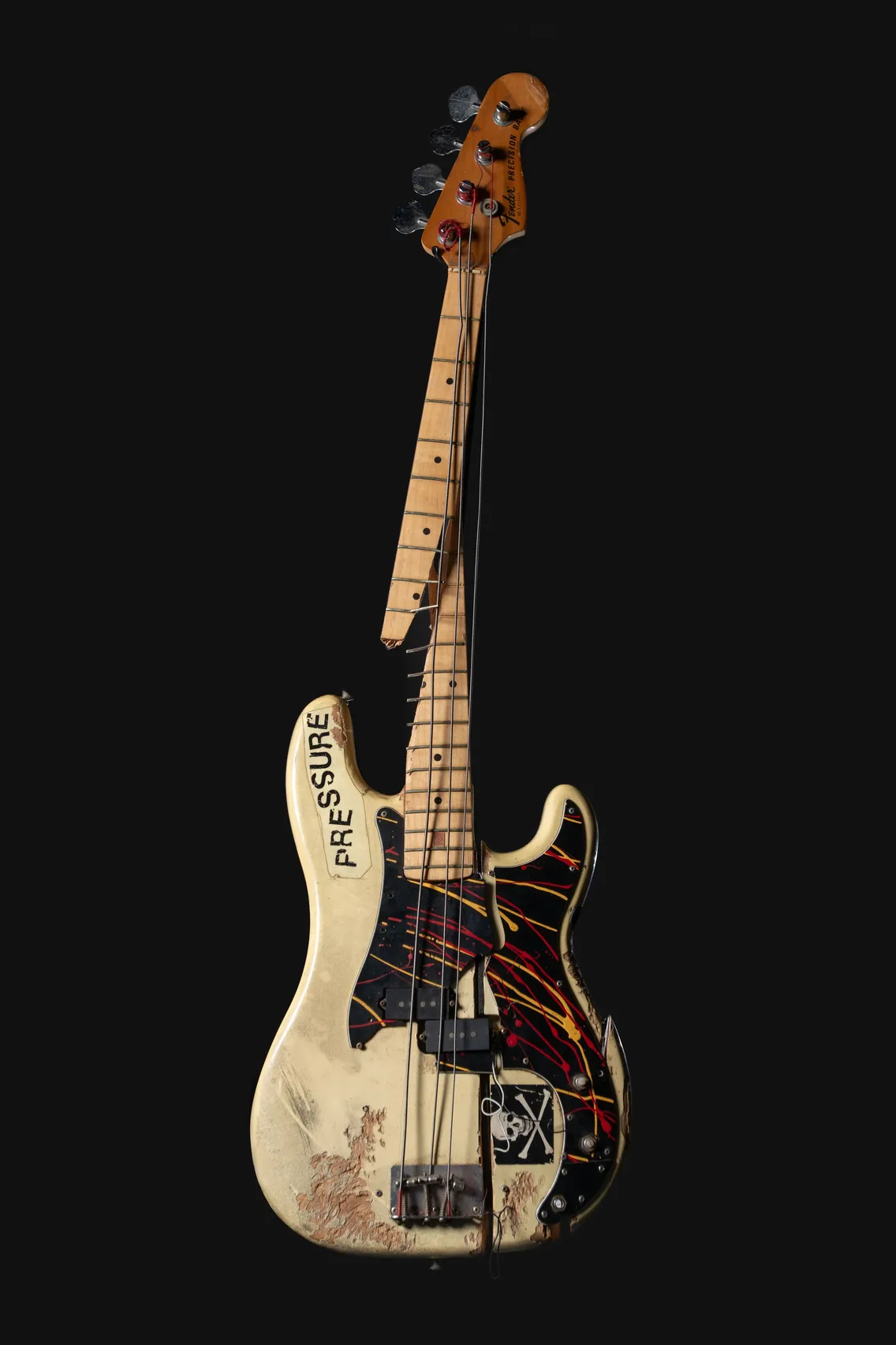 A damaged electric bass guitar with a broken neck and worn body, decorated with stickers and the word "PRESSURE" on the front, against a black background.