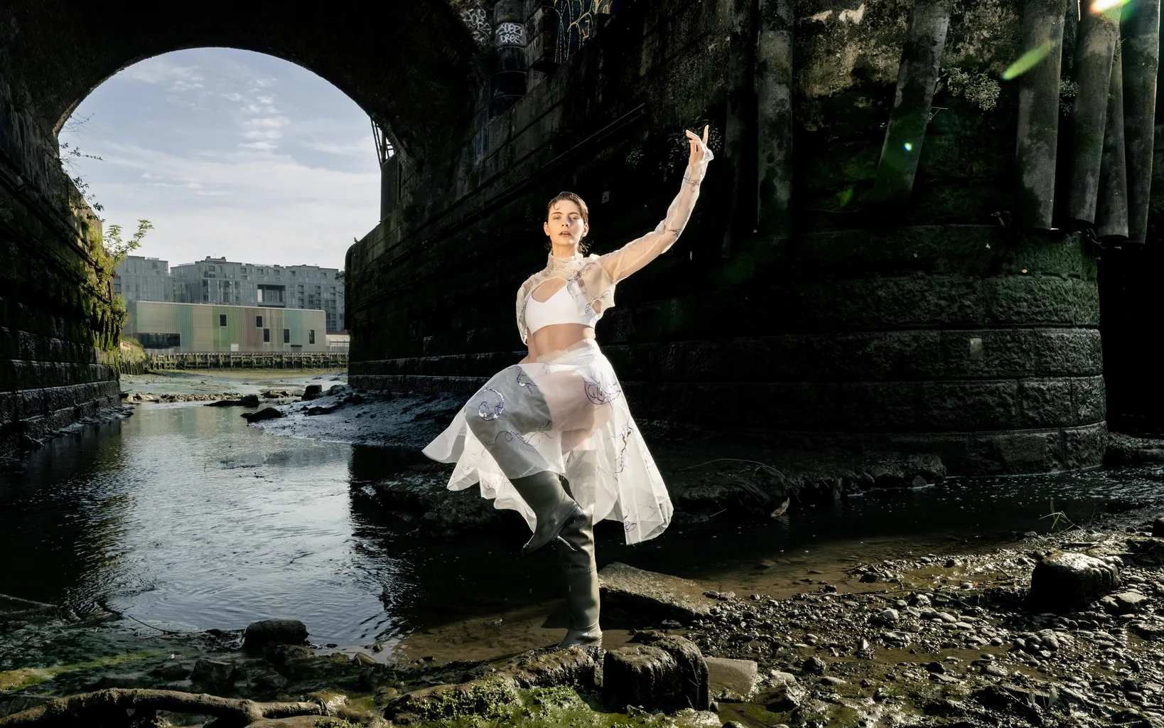 A person wearing wellington boots on the foreshore, posing with their leg and arm up