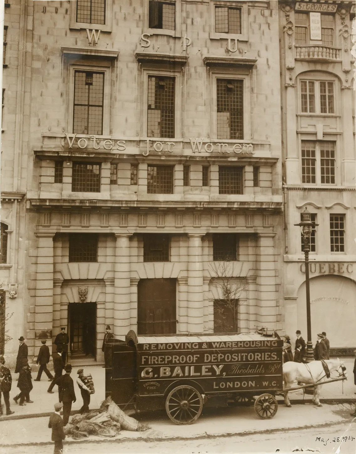 Black and white historical photo showing a building with "votes for women" banners and a horse-drawn wagon marked "c. bailey" in front, dated may 28, 1914.