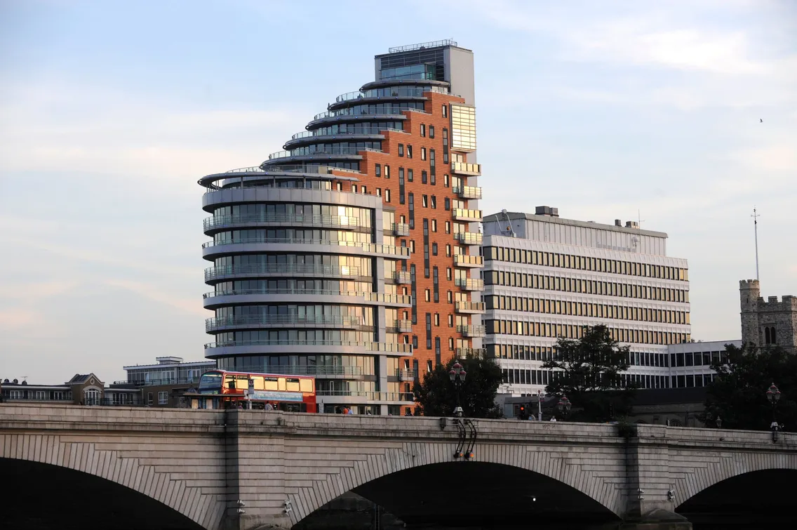 A modern, curved glass and brick building stands next to an older office block behind a stone bridge with a red double-decker bus crossing it.