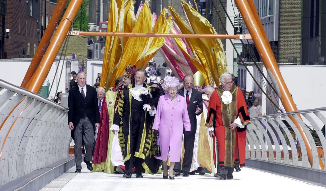 A group of formally dressed people, including a woman in a lavender coat and hat, walk across a bridge decorated with large yellow and pink fabric banners.